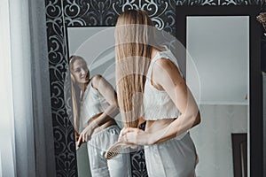 How take care of long hair. Back view of young natural blonde woman in white home clothes combing her hair and looking