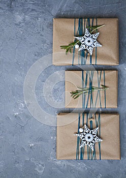 Hree Christmas gifts in craft paper wrapped in blue thread, with white snowflakes and rosemary branches on a gray concrete