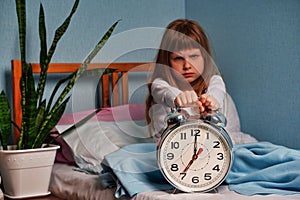 Huge alarm clock in the hands of a angry sad child girl in the bedroom. 7 o& x27;clock in the morning, stress.