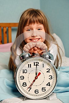 Huge alarm clock in the hands of a happy smile child girl in the bedroom. 7 o'clock in the morning.