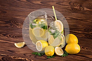 A huge jug and glass full of fresh summer lemonade with yellow, ripe, and juicy lemon and fresh green mint on a dark wooden table.