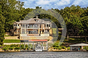 Huge lake house with two layers of deck toward water with big motor yacht parked in private dock and beautiful trees in background