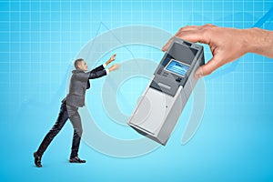 Huge man`s hand giving miniature ATM to little businessman on light blue background with slightly visible business