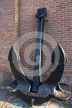 Huge vintage black metal anchor with two large pointed paws standing on the ground, propped up and attached to a red brick wall