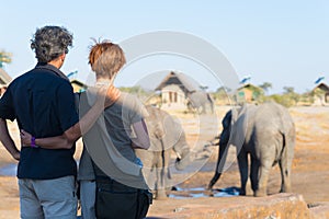 Hugging couple looking at elephant herd drinking from waterhole. Adventure and wildlife safari in Africa. People traveling concept
