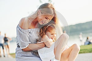 Hugs and tickles. Photo of young mother and her daughter having good time on the green grass with lake at background