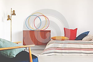 Hula hoops on a cupboard next to a bed with colorful pillows in a retro bedroom interior