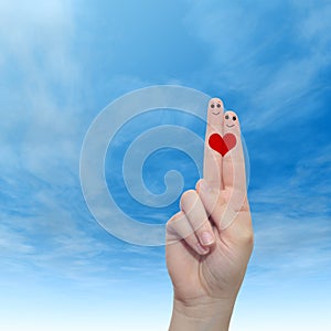 Human or female hands with two fingers painted with a red heart and smiley faces over cloud blue sky