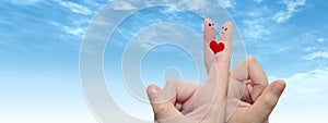 Human or female hands with two fingers painted with a red heart and smiley faces over cloud blue sky