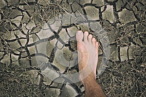 Human foot on dried cracked lifeless earth. Man`s barefoot on dry soil with dead plants. Barren land because of ecological