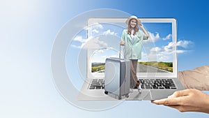 A human hand holding a laptop with an Asian girl in a hat and a suitcase with a hill view and blue sky on the laptop screen