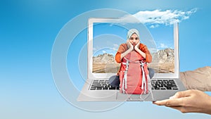 A human hand holding a laptop with an Asian girl in a scarf with a backpack sitting with a rock cliff view and blue sky on the