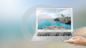 A human hand holding a laptop with a screen view of the sea cliff with an ocean view and blue sky