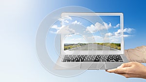 A human hand holding a laptop with a screen view of the street with green hills and a landscape view with blue sky