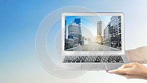 A human hand holding a laptop with a screen view of the street and a skyscraper with a city view and blue sky