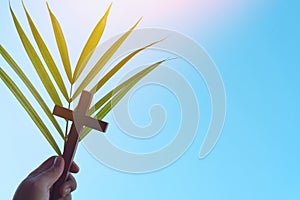 Human hand holding palm leaves with wooden crucifix cross in clear blue sky. Palm sunday background.