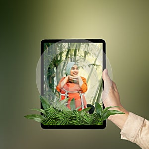 A human hand holding tablet with a screen view of an Asian girl in a scarf sitting with a backpack in the rainforest with trees