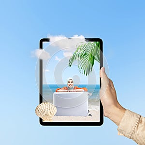 A human hand holding tablet with a screen view of an Asian girl in a scarf sitting with a suitcase on the beach with an ocean view