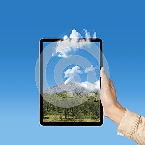 Human hand holding tablet with a screen view of the trees with mountain view and blue sky