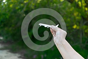 Human hand holding white airplane model in front of green tree background. Transportation trip concept