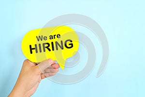 Human hand holding yellow speech bubble with written We are Hiring. Recruitment and job hiring concept.