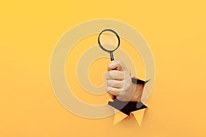 A human hand holds a magnifying glass through a torn hole in a paper yellow wall. Search information concept