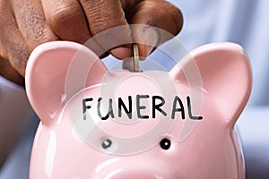 Human Hand Inserting Coin In Piggy Bank With Funeral Text