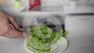 Human hand putting bunch of green grapes on white plate on kitchen table. Healthy and fresh food, good source of