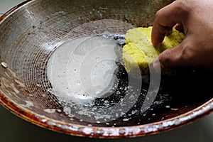 Human hand scrubbing or cleaning a burnt, scorched and greasy pan or wok on kitchen sink.
