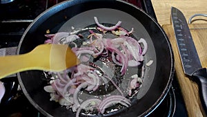 Human hand stirring fried onion in frying pan in domestic kitchen for cooking food on kitchen stove.