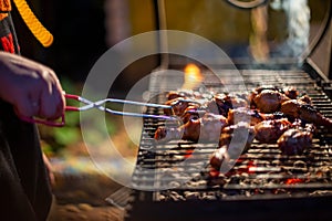 a human hand turns chicken drumsticks on a barbecue grill with grilling tongs. cooking food on an open fire in the
