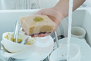 Human hand wash white dinnerware with dish soap on sponge under tap water. Pile of kitchen utensils in sink closeup.