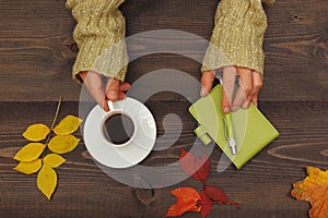 Human hands with a cup of coffee at a wooden table with notebook and pen and autumn leaves