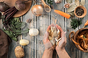 Human hands hold handful of peeled potato peel, flat lay. Mix of fresh vegetables on wooden table. Zero waste concept.