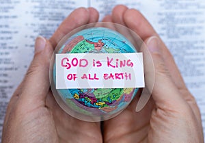 Human hands hold handwritten verse quote, God is King of all earth upon world globe and open Holy Bible Book