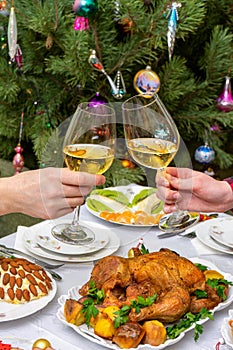 Human hands holding glasses with sparkling wine against beautiful decorated Christmas tree and served table with tasty dishes. New