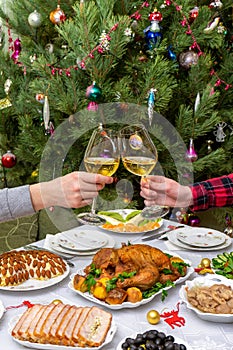 Human hands holding glasses with sparkling wine against beautiful decorated Christmas tree and served table with tasty dishes. New