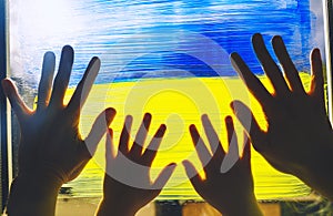 Human hands of parent and child touch painting yellow-blue flag of Ukraine on window. Adult and kids hands on image of flag of