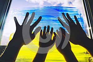 Human hands of parent and child touch painting yellow-blue flag of Ukraine on window. Adult and kids hands on image of flag of