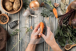 Human hands peel ripe carrot with vegetable peeler, flat lay. Fresh vegetables on wooden table. Zero waste concept.