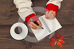Human hands with a pen and notebook at a wooden table with cup of espresso and autumn leaves