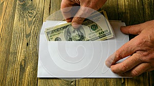 Human hands put two one hundred dollar bills in cash into white paper envelope that lies on brown wooden table. Concept of saving