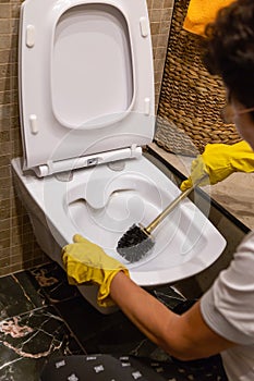 Human hands in rubber gloves cleaning the toilet bowl with brush and rag in a bathroom, vertical