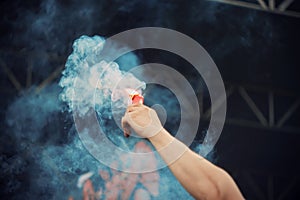 A human holds a red signal fire in his hand, which emits blue smoke