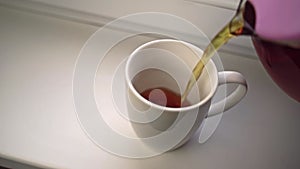 Human pours fragrant tea from glass teapot into white ceramic mug.