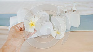 Human`s hand holding white blossom plumeria flower with blurred bath accessories on bed background
