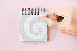 Human`s hand holding white thumbtack to mark date on blurred white calendar with sweet pink background