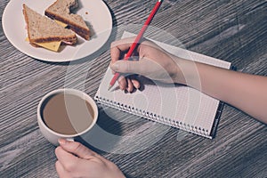 A human`s hand writing in a notebook. Break at work. Background of a hand holding a cup of hot coffee, tasty cheese sandwich. Top