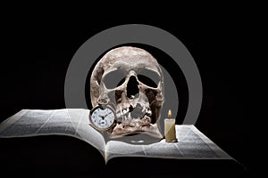 Human skull on old open book with burning candle and vintage clock on black background under beam of light