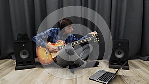 Human tuning electric guitar. Man in headphones  tunes guitar using notebook while sitting on floor at living room. Young professi